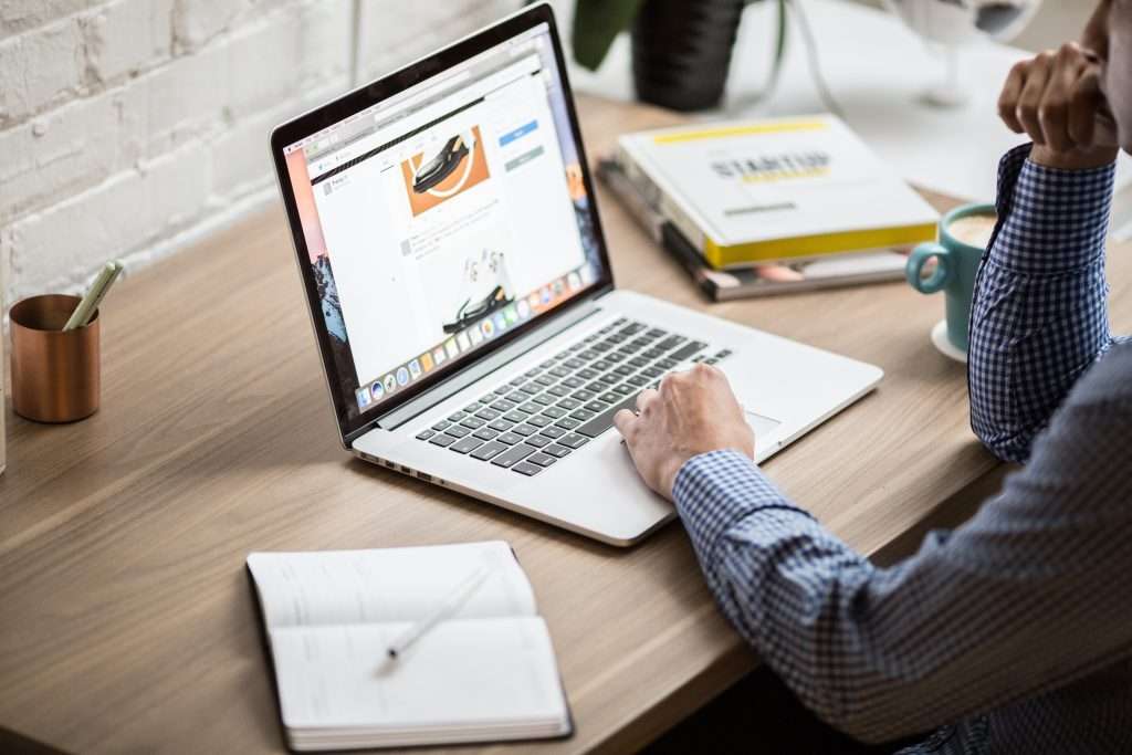 A man in blue checkered shirt sitting at a Mac computer with his left hand on the mousepad while his right hand holds his chin