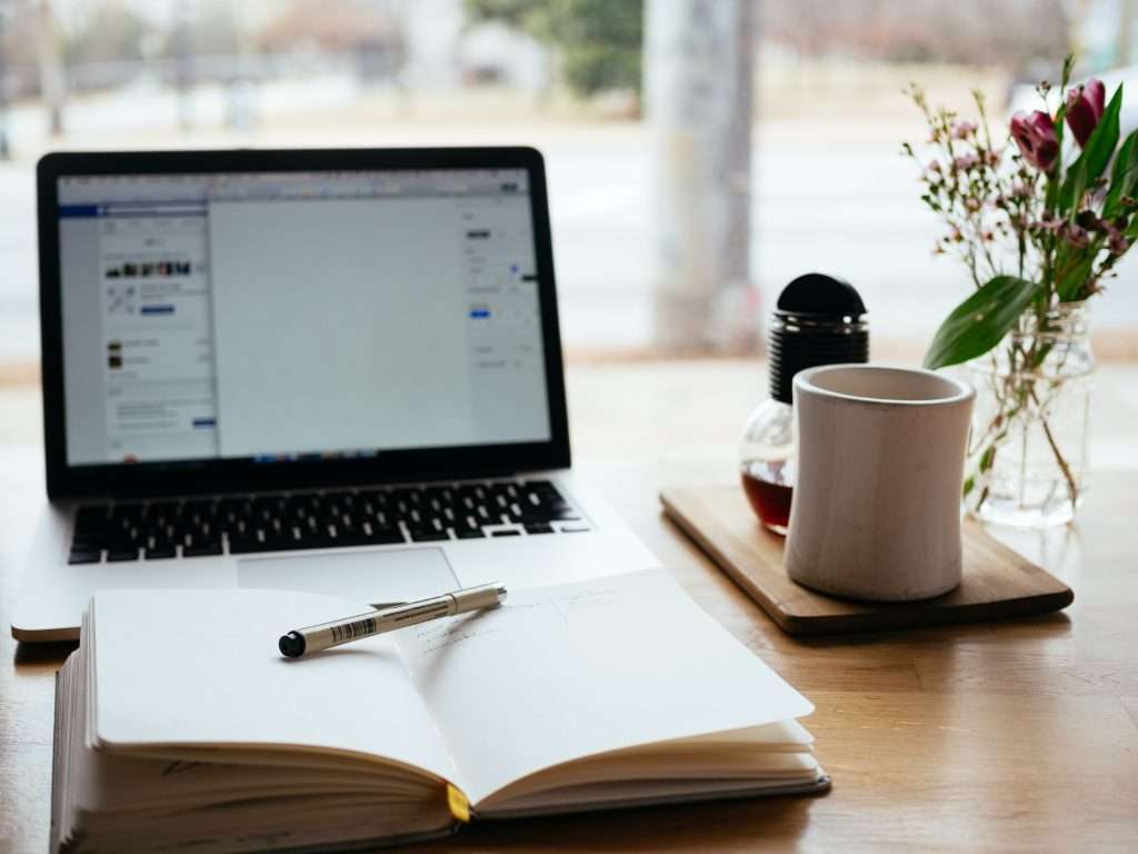 Mac on a wooden table with a cup of coffee and small flowers in a glass vase off to the right side, with a notebook with pen on top in front of the computer.