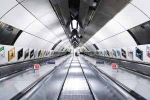 Looking down escalators of a London tube station with posted ads on the sides of the walls