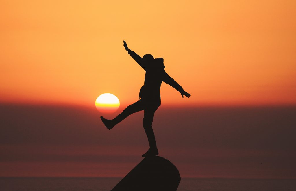 A silhouetted person standing on one leg on a rock with the sunset in the background