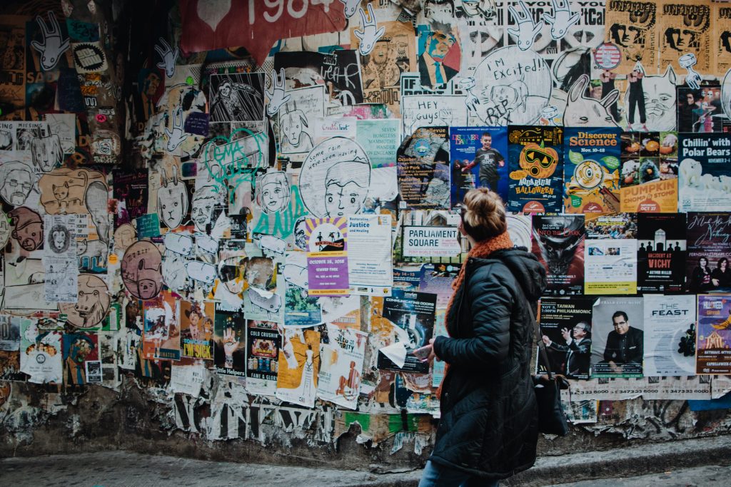 A woman with a black coat on looks away from the camera at a wall full of taped paper advertisements taped