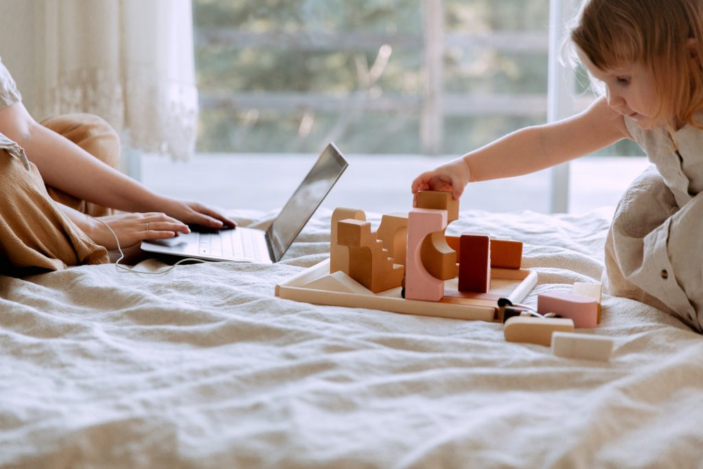 An adult woman working on their bed with a computer open, as a child sits near playing with wooden blocks.
