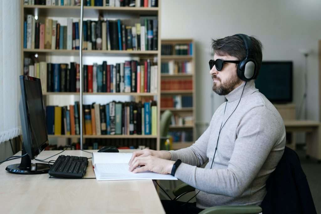 A blind man wearing a headset sits in front of a desktop computer with a Braille book in front of him