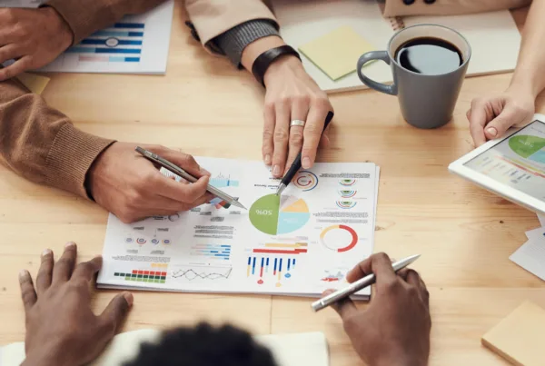A group of 4 people sitting around a table with only their hands showing as they point at papers with graphs and charts on them
