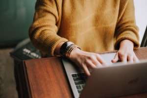 Person, wearing a mustard yellow sweater, sits and types on a Mac computer