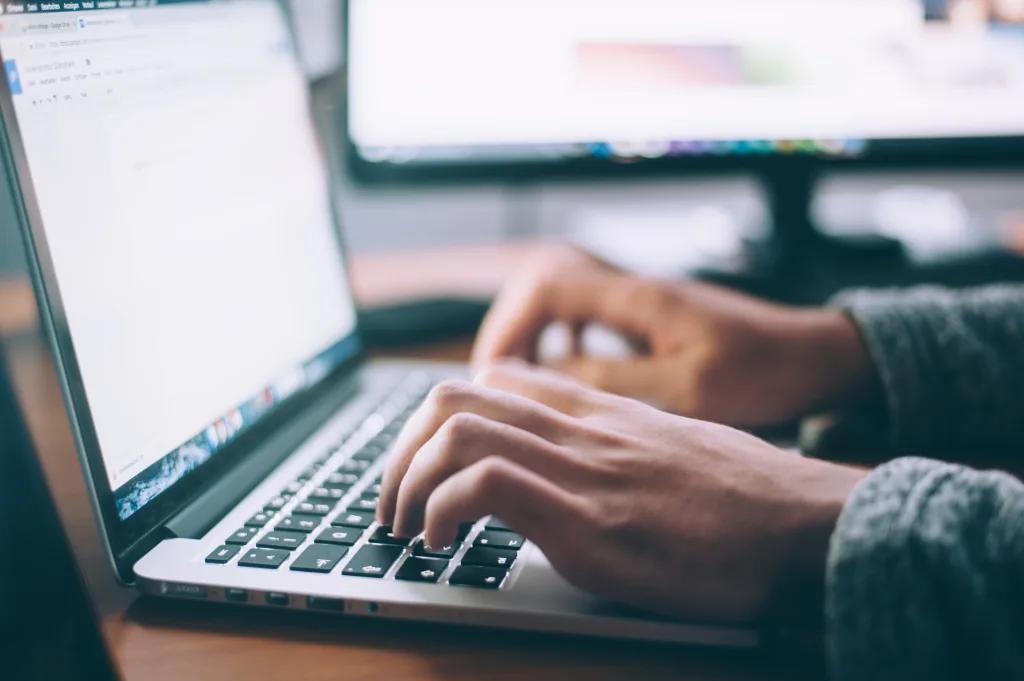 Hands of a person (who is wearing a denim shirt) typing on a Mac computer with a bright screen