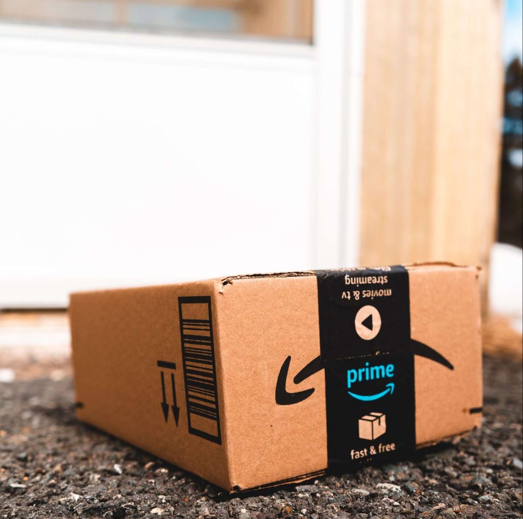 An Amazon Prime cardboard box sitting on asphalt ground outside of a white door.