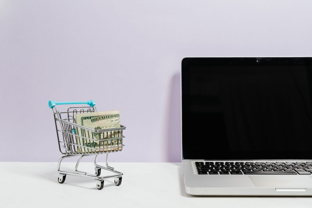 Tiny shopping cart with paper money tucked inside it with a blank screened Mac to the right